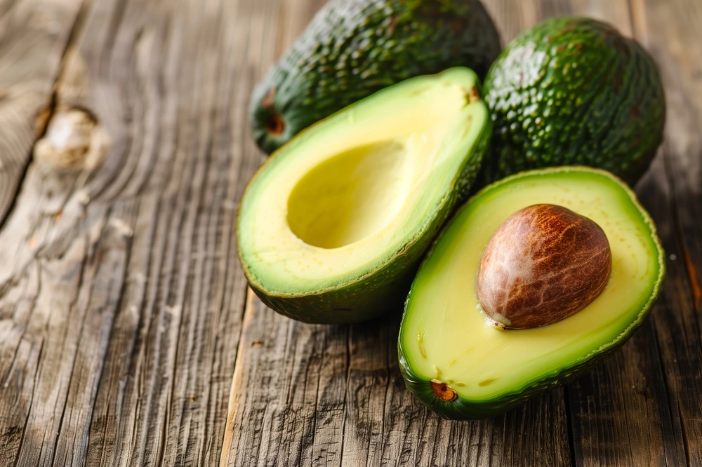 Three avocados on a wooden table - one of which is cut in half revealing a whole seed inside.