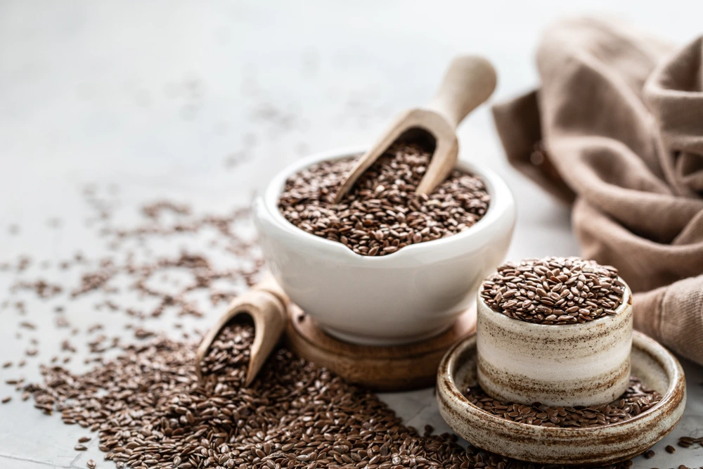 Flaxseeds filling a white dish, a small white cup and two scoops with flaxseeds all over the floor.