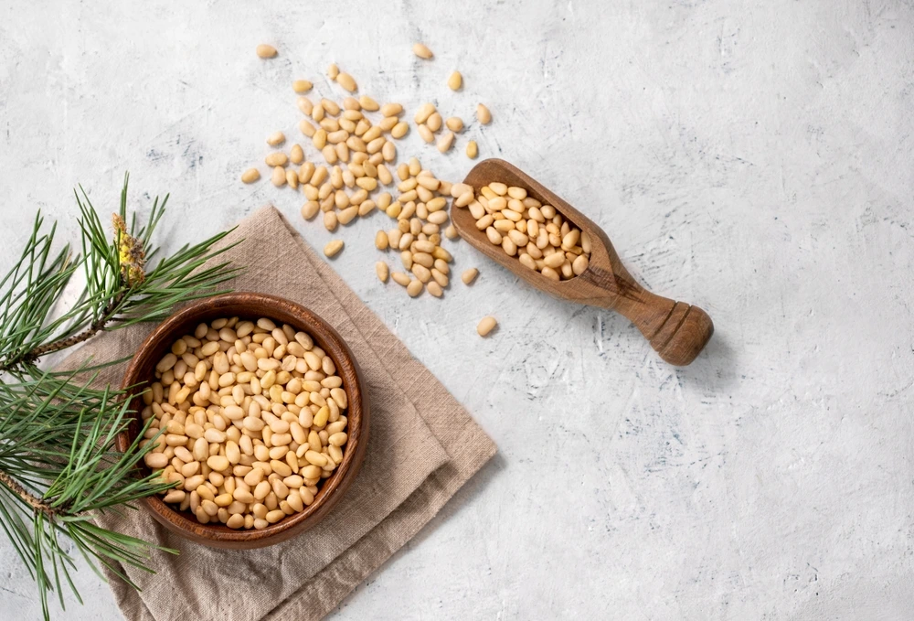 Pine nuts fill a wooden bowl on a brown cloth next to some pine. There is also a serving scoop with pine nuts in it.