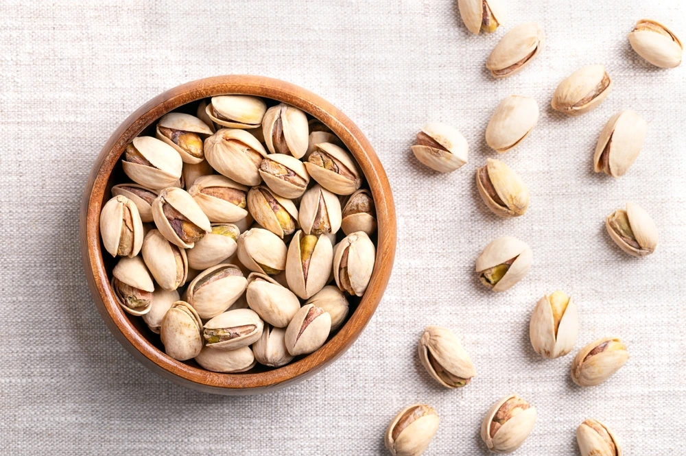 Pistachio nuts in a wooden bowl with a white background and some nuts beside the bowl.