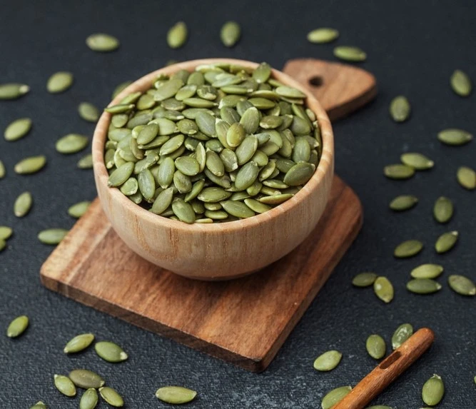 Pumpkin seeds filling a wooden bowl on a wooden board with some seeds scattered aroudn on a black table.